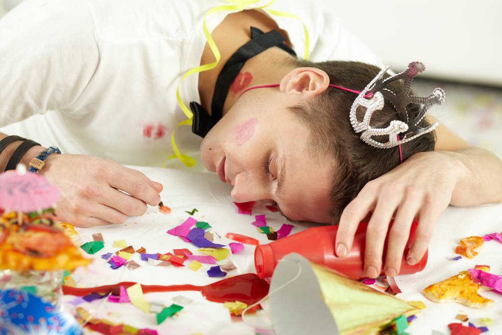 Young man in party hat sleeping on table among confetti and remnants of food