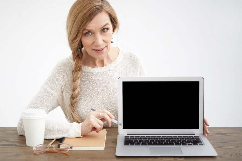 Attractive European woman pensioner with long braid sitting at her workplace with coffee, glasses and copybook, holding pen and pointing it at blank copyspace screen of generic portable computer
