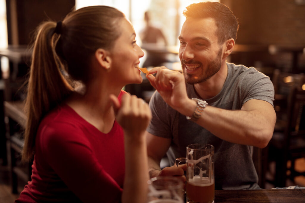 Happy man feeding his girlfriend with nacho chips while drinking beer together in a pub.